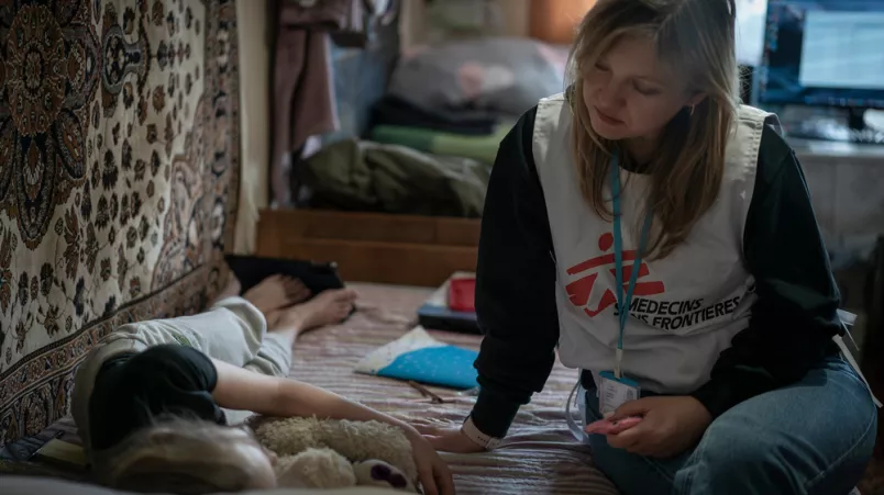 Ein Kind liegt im Bett und eine Mitarbeiterin sitz neben ihr und redet mit ihr.