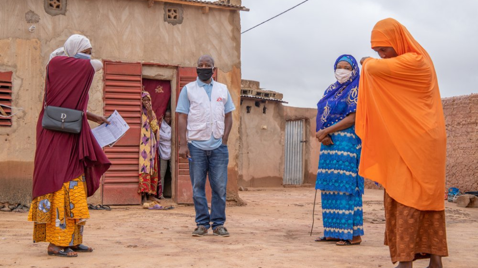 Drei Frauen und ein Mann stehen in einem Kreis, eine Frau zeigt der anderen wie sie korrekt in die Armbeuge husten muss, als Vorsichtsmaßnahme gegen das Coronavirus