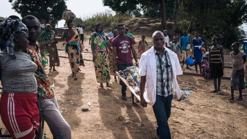 Zwei Männer tragen eine Tragbahre mit einer Patientin, darum stehen Menschen und schauen ihnen zu