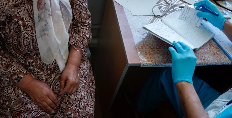 Man sieht eine Person im Kleid, das Gesicht wird nicht gezeigt und eine Person in ärztlicher Kleidung, auch hier ist das Gesicht nicht zu sehen. Beide sitzen an einen Tisch.