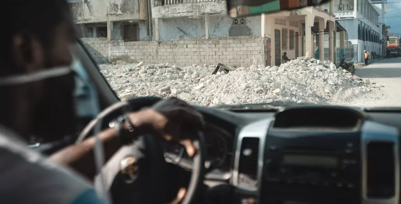 Einer unserer Mitarbeiter sitzt in einem Auto. Im Hintergrund ist die Zerstörung durch ein Erdbeben sichtbar.