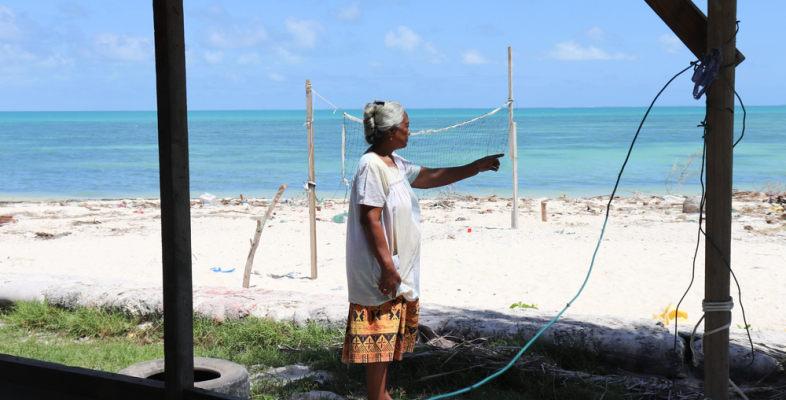 Eine Frau zeigt, wie sich der Strand vor ihrem Haus verschmalert. Am Horizont ist das Meer, das ihrem Haus immer näher rückt.