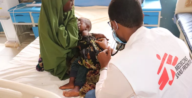 Foto von einer Frau mit ihrem Baby auf dem Arm, welches von einem Arzt untersucht wird. Der Arzt ist von hinten sichtbar und das Ärzte ohne Grenzen-Logo ist auf seinem Gilet aufgedruckt