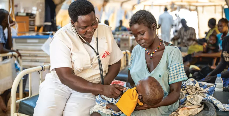 Eine Mitarbeitende untersucht ein Kind in den Armen seiner Mutter