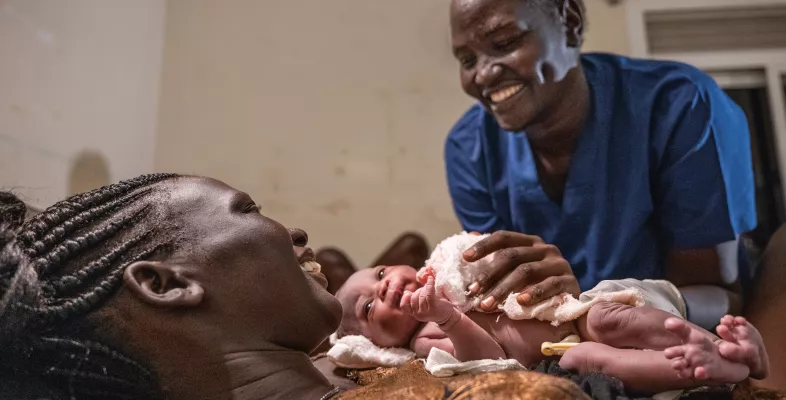 Eine Frau hält ein Baby in den Armen, am Bett steht eine Hebamme