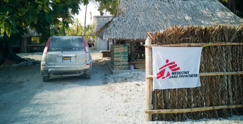 Parkendes Auto neben Gebäuden mit Strohdach, davor ein Zaun