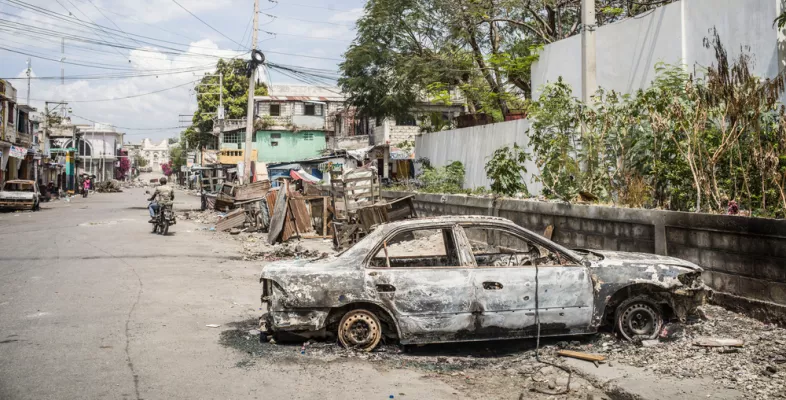 Auf der Straße steht ein ausgebranntes Auto, im Hintergrund fährt jemand auf einem Moped.