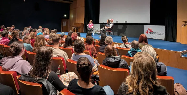 Blick aus dem Publikum auf drei Personen auf der Bühne während eines Live-Gesprächs. Der Saal wirkt voll.