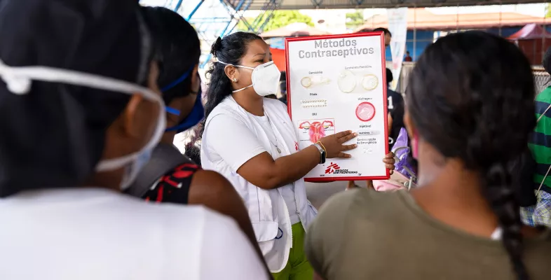 Eine Frau mit Maske spricht vor Anderen, ein Plakat zu Kontrazeptiva in der Hand