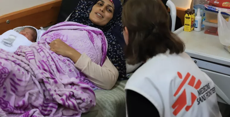 Eine Frau liegt mit Neugeborenem im Bett, eine Mitarbeiterin spricht mit ihr.