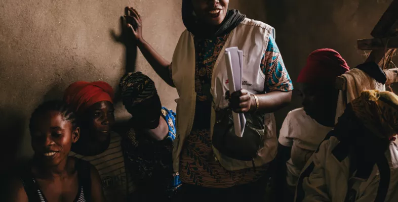 Eine Frau mit Weste spricht in einem schwach beleuchteten Raum zu mehreren sitzenden Frauen und hält dabei einige Papiere in der Hand.