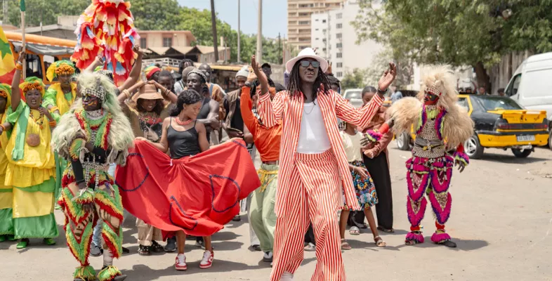 Eine Gruppe von Menschen tanzt und feiert auf einer Straße in Dakar. Vorne singt ein Mann im rot-weiß gestreiften Anzug.