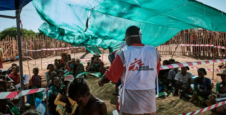 Ein Ärzte ohne Grenzen Mitarbeiter steht vor Patient*innen, die zum Schutz vor der Sonne unter einer aufgespannten Plane sitzen.