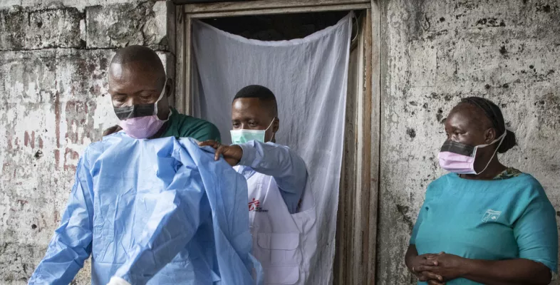 Ein medizinischer Mitarbeiter bekommt einen OP-Kittel angezogen. Eine weitere Mitarbeitern sieht zu