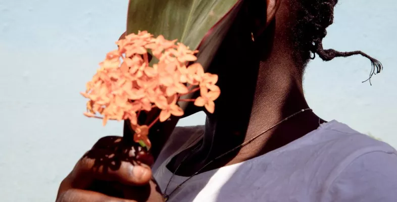 Person die sich ein grünes Pflanzenblatt und eine orangene Blume vor das Gesicht hält um nicht erkannt zu werden 