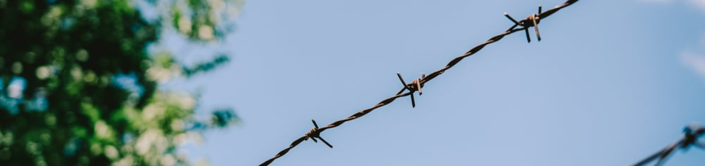 Gewalt gegen Geflüchtete und Migrant*innen Ein Zaun mit Stacheldraht vor blauem Himmel