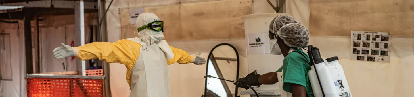 Eine Person in Schutzkleidung wird in einem Zelt von einem Mitarbeitenden mit Desinfektionsspray dekontaminiert.
