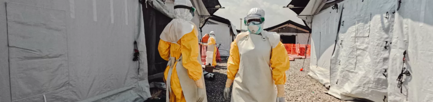 Mehrere Mitarbeitenden in Schutzanzügen stehen im freien vor einem Ebola Behandlungszentrum.