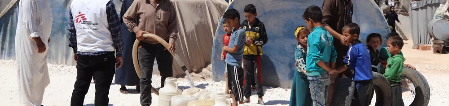 Mitarbeiter von Ärzte ohne Grenzen befüllen Wasserkanister, daneben eine Gruppe von Kindern