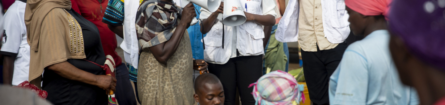 Ärzte ohne Grenzen Aufklärungsarbeit zum Thema Cholera. Eine Community Health Mitarbeiterin klärt mit einem Megaphon auf dem Buterere-Markt in Bujumbura über Cholera auf. Um sie herum sind mehrere Mitarbeiter sowie weitere Personen versammelt und hören zu.