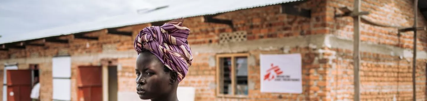 Eine Frau steht mit einem Kind auf dem Rücken vor einem Backsteingebäude mit Logo von Ärzte ohne Grenzen