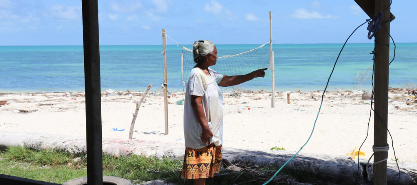 Eine Frau zeigt, wie sich der Strand vor ihrem Haus verschmalert. Am Horizont ist das Meer, das ihrem Haus immer näher rückt.