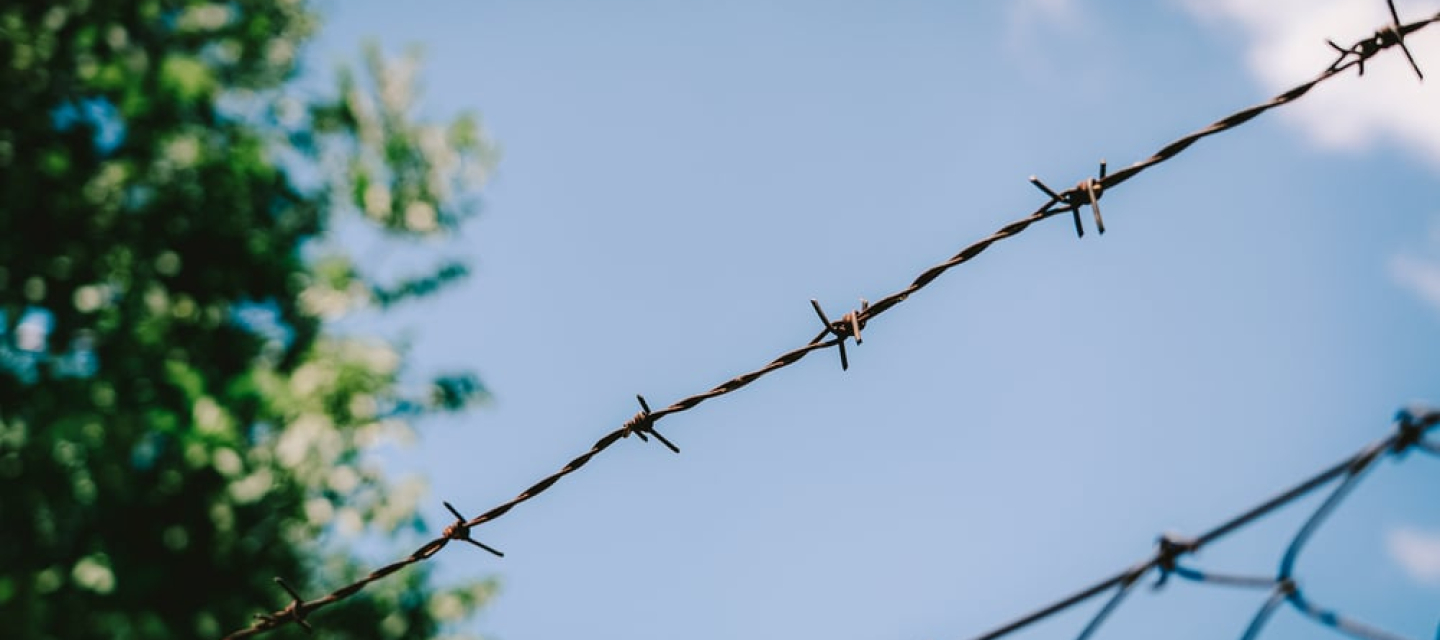 Gewalt gegen Geflüchtete und Migrant*innen Ein Zaun mit Stacheldraht vor blauem Himmel