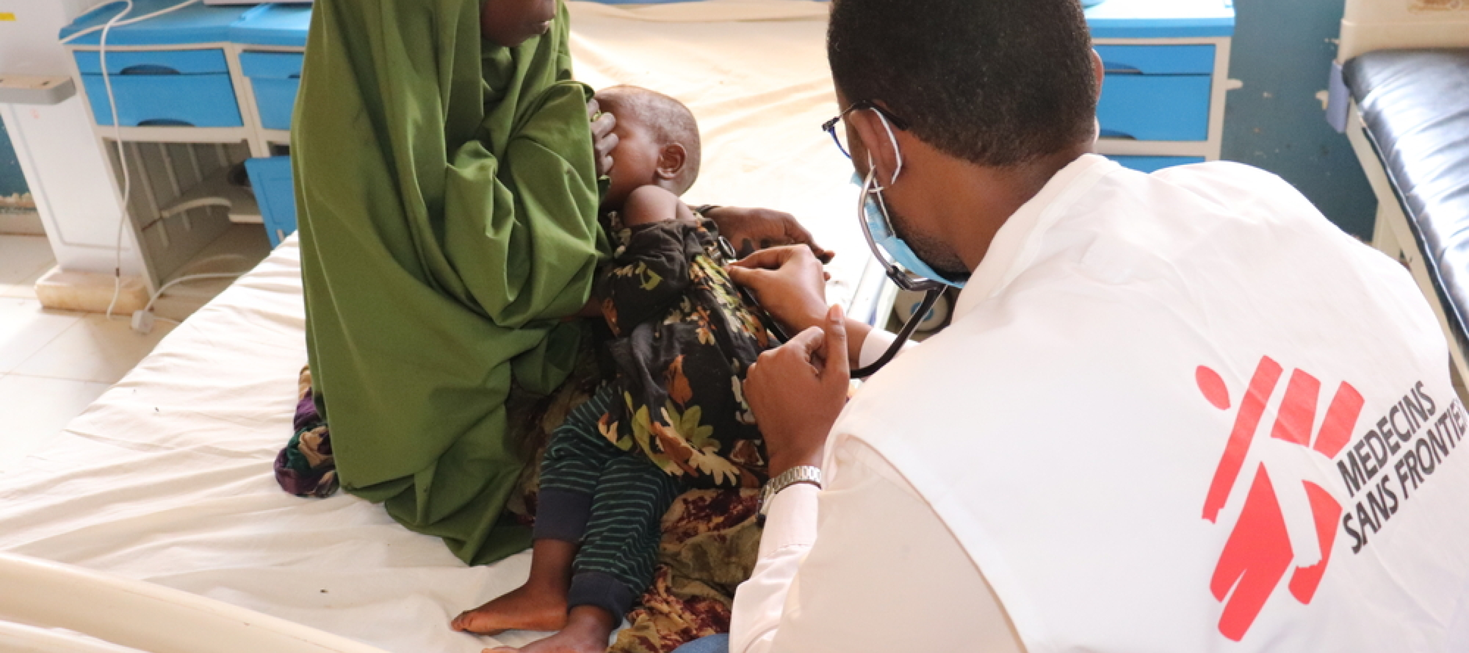 Der Arzt Ahmed Ilyas untersucht das Baby Saidya im Bay Regional Hospital in Baidoa, Somalia. Foto von einer Frau mit ihrem Baby auf dem Arm, welches von einem Arzt untersucht wird. Der Arzt ist von hinten sichtbar und das Ärzte ohne Grenzen-Logo ist auf seinem Gilet aufgedruckt