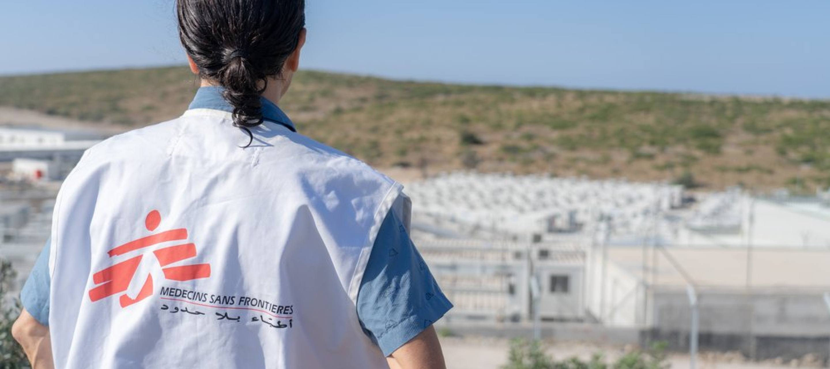 Eine Person von hinten fotografiert, mit einer Ärzte ohne Grenzen Weste die auf ein entferntes Camp schaut