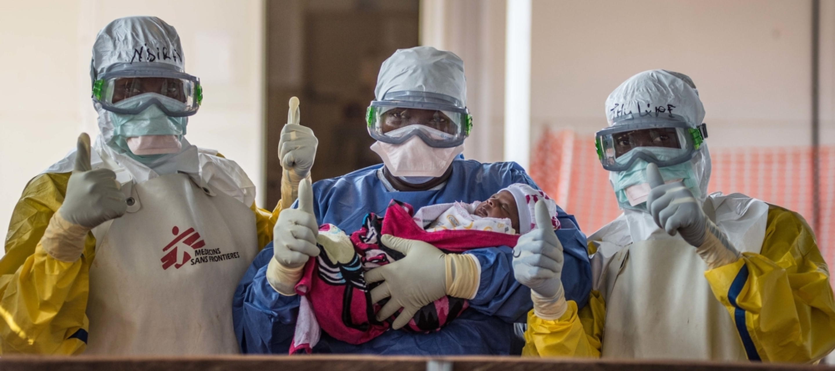 Ein an Ebola erkranktes Baby kann das Behandlungszentrum in Conakry, Guinea, verlassen. Es hat den Kampf gegen das Virus gewonnen. Drei Ärzte*innen und ein Baby