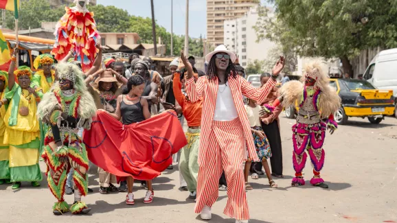 Eine Gruppe von Menschen tanzt und feiert auf einer Straße in Dakar. Vorne singt ein Mann im rot-weiß gestreiften Anzug.
