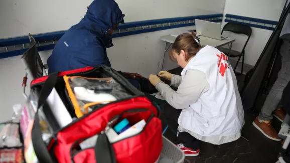 Eine Mitarbeiterin von Ärzte ohne Grenzen versorgt mit Handschuhen das Knie einer sitzenden Person, während im Vordergrund eine geöffnete rote Notfalltasche steht.