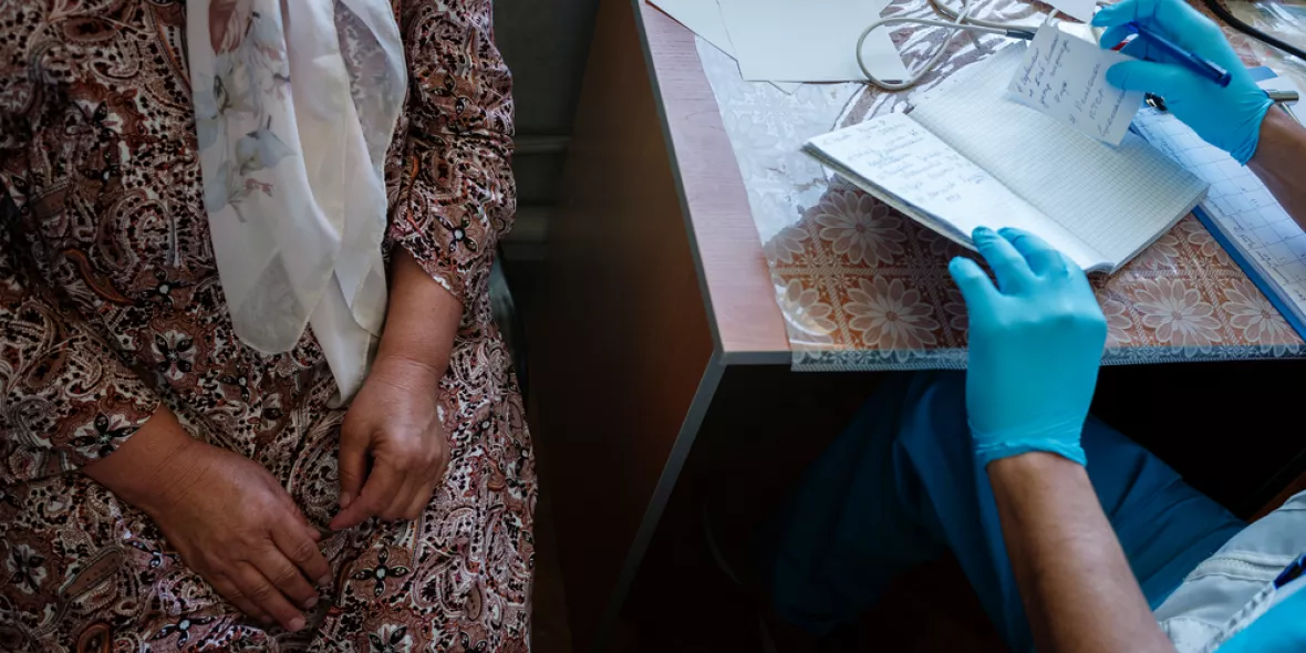 Man sieht eine Person im Kleid, das Gesicht wird nicht gezeigt und eine Person in ärztlicher Kleidung, auch hier ist das Gesicht nicht zu sehen. Beide sitzen an einen Tisch.