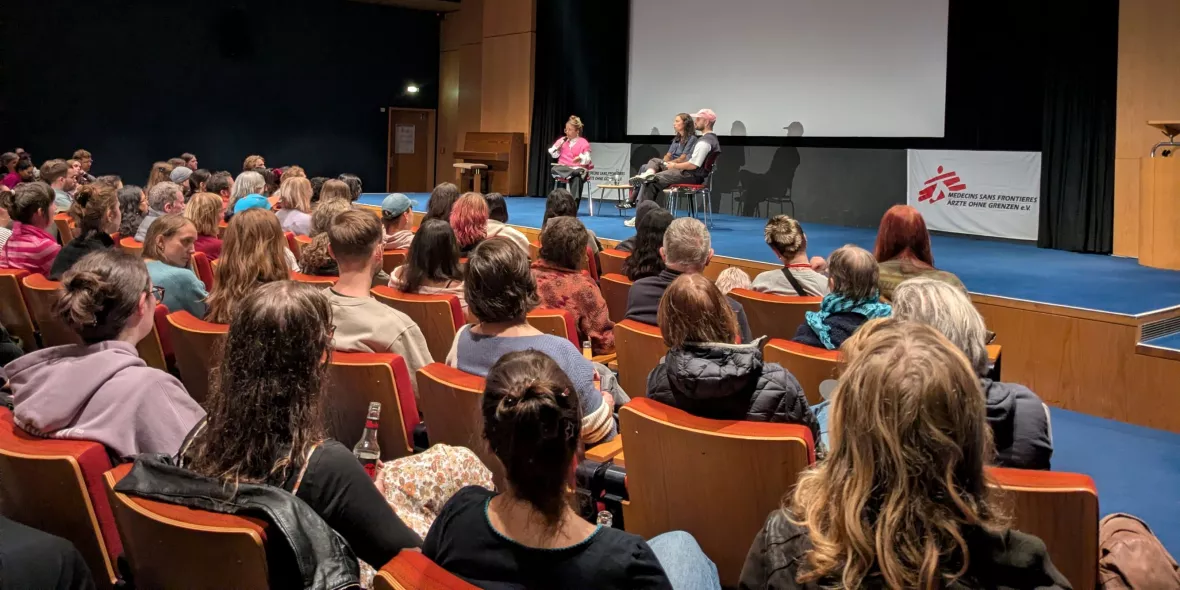 Blick aus dem Publikum auf drei Personen auf der Bühne während eines Live-Gesprächs. Der Saal wirkt voll.