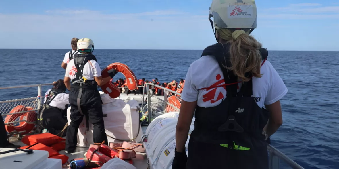Eine Person steht auf einem Schiff und schaut mit zwei andere Personen geradeaus auf ein kleines Boot mit vielen geflüchteten.