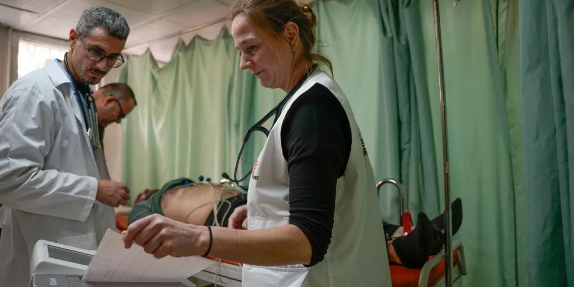 Eine unserer Mitarbeiterinnen hält einen EKG-Ausdruck, während neben ihr ein Mitarbeiter und im Hintergrund ein Patient auf einer Liege zu sehen sind.