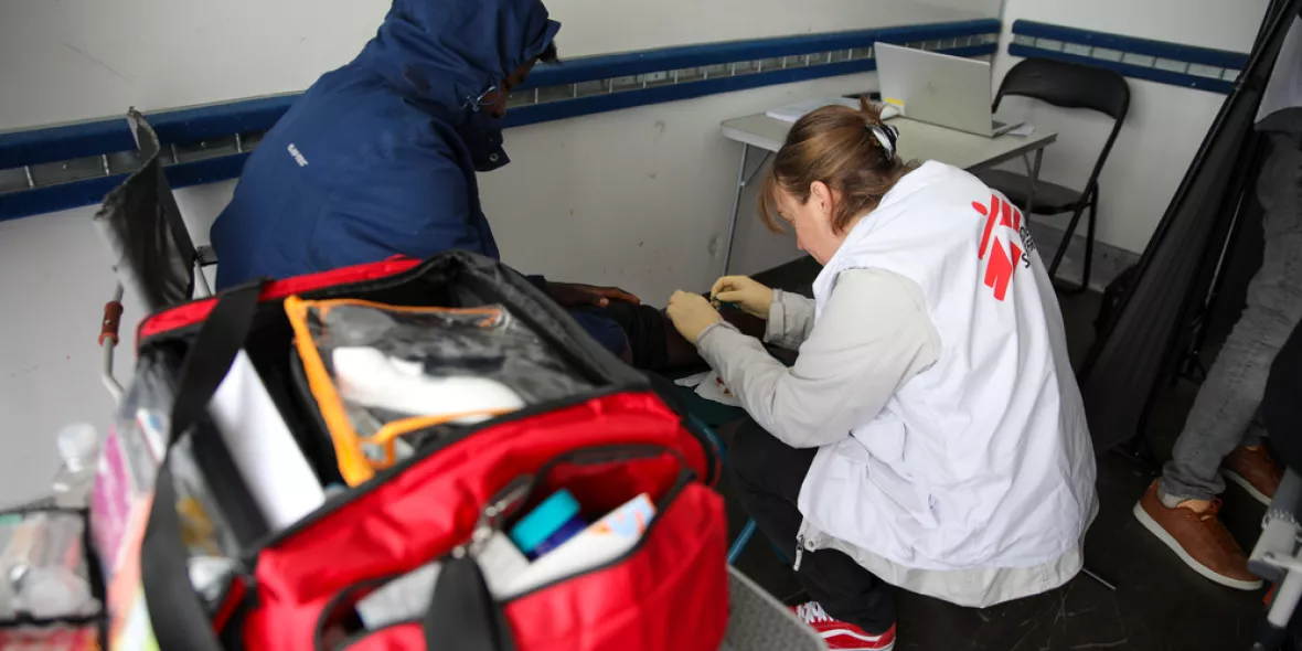 Eine Mitarbeiterin von Ärzte ohne Grenzen versorgt mit Handschuhen das Knie einer sitzenden Person, während im Vordergrund eine geöffnete rote Notfalltasche steht.