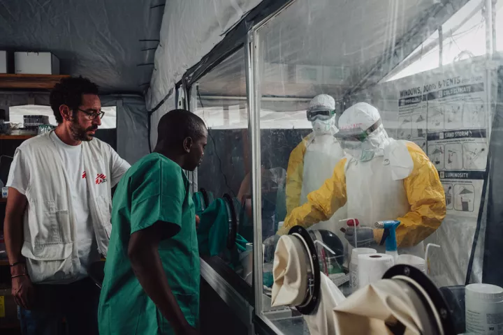 Zwei Personen stehen in Schutzanzügen hinter einer Glasscheibe in einem Labor und interagieren mit zwei medizinischen Mitarbeitern.