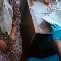 Man sieht eine Person im Kleid, das Gesicht wird nicht gezeigt und eine Person in ärztlicher Kleidung, auch hier ist das Gesicht nicht zu sehen. Beide sitzen an einen Tisch.