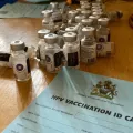 Auf einem Tisch stehen HPV Impfdosen und ein Impfzertifikat.