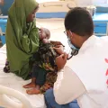 Foto von einer Frau mit ihrem Baby auf dem Arm, welches von einem Arzt untersucht wird. Der Arzt ist von hinten sichtbar und das Ärzte ohne Grenzen-Logo ist auf seinem Gilet aufgedruckt
