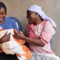 Zwei Frauen sitzen auf einer Bank. Eine hält ein Baby im Arm.