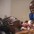 Eine Frau hält ein Baby in den Armen, am Bett steht eine Hebamme