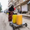Eine Frau transportiert zwei Wasserkanister auf einem Karren durch eine Straße.