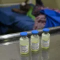 Drei Impfampullen stehen auf einem Tisch in einem Krankenhaus. Im Hintergrund ist eine Patientin zu sehen.