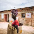 Eine Frau steht mit einem Kind auf dem Rücken vor einem Backsteingebäude mit Logo von Ärzte ohne Grenzen