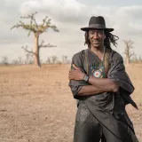 Ein Mann mit Hut steht mit verschränkten Armen in einer trockenen Landschaft mit einzelnen Bäumen im Hintergrund.
